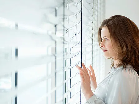 Una mujer mira por la ventana entre las lamas de una persiana veneciana